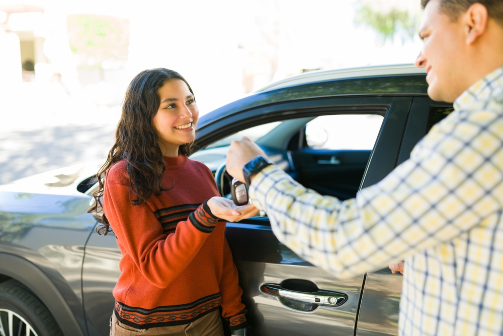 FatherHappilyPassingCarKeysToHisTeenageDaughterAnd Father handing car keys to his teenage driver before getting behind the wheel.